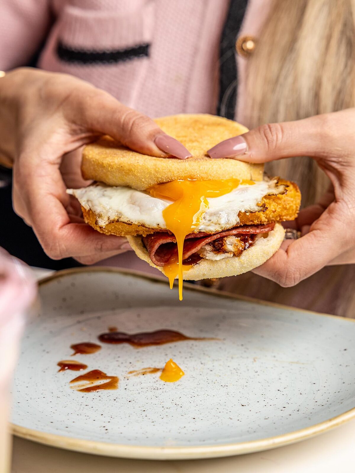 Breakfast muffin with fried egg, bacon and hash brown, the yolk running out as it is pulled apart