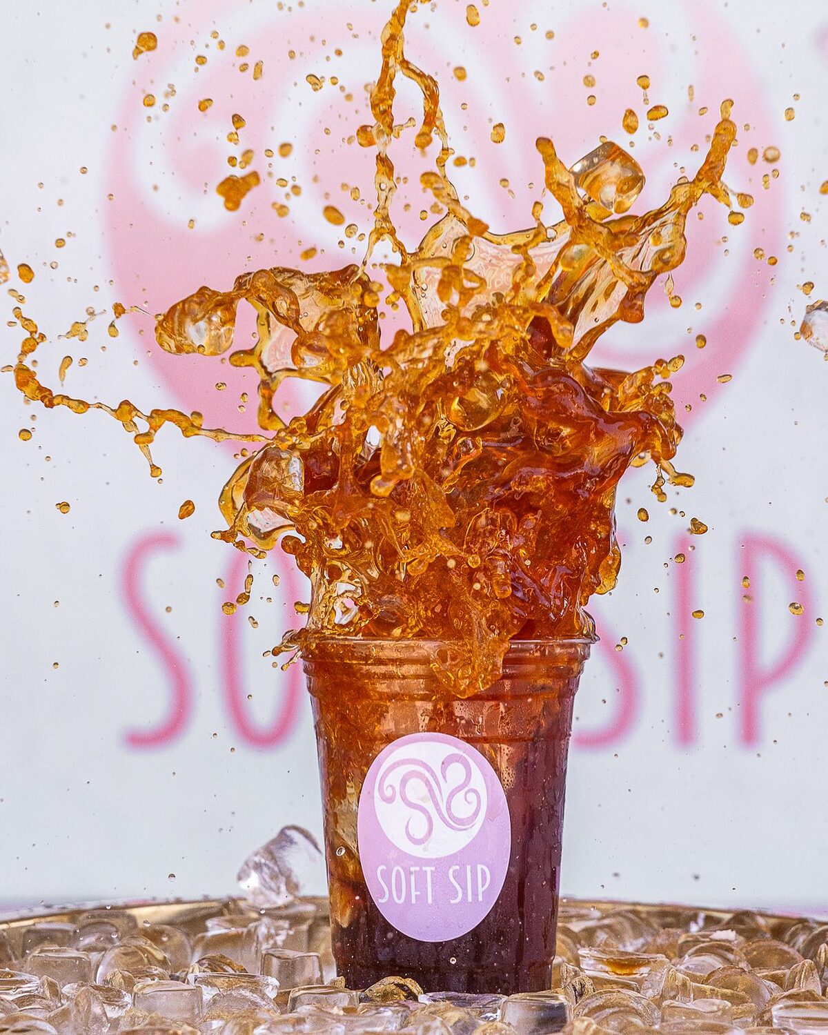 Iced coffee splashing out of a Soft Sip cup in front of the pink logo wall