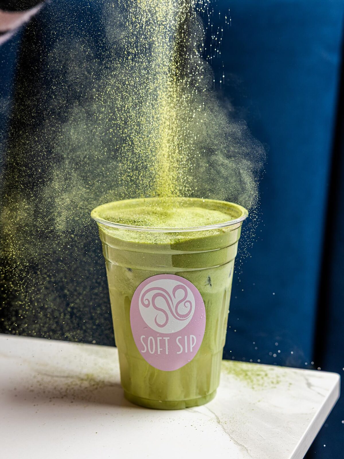 A cloud of matcha powder above an iced matcha latte in a Soft Sip cup