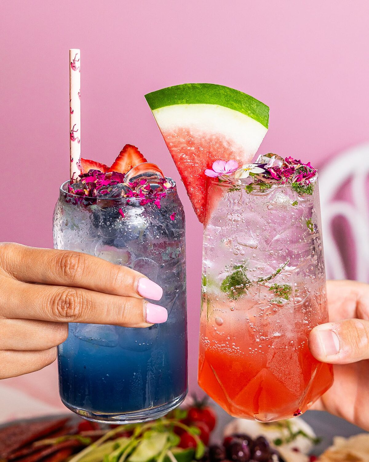 Two iced mocktails with watermelon, strawberry and rose petals against a pink wall